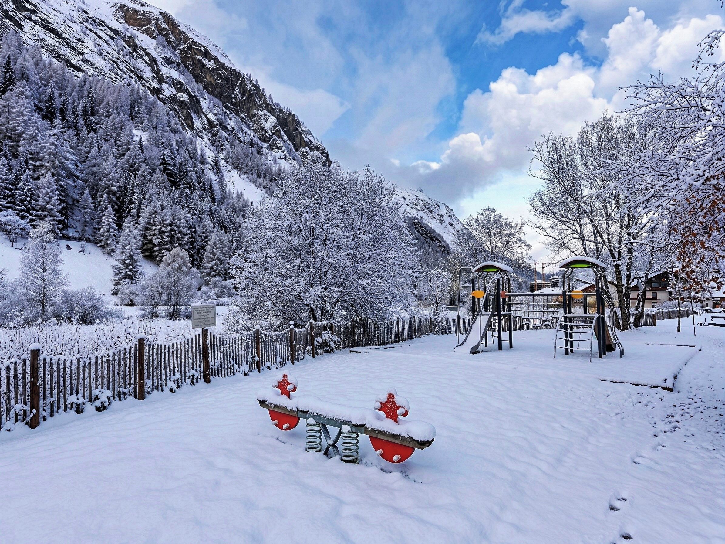 A snow-covered playground with slides and swings, surrounded by snow-laden trees and mountains.