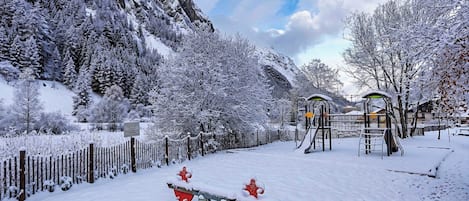 A snow-covered playground with slides and swings, surrounded by snow-laden trees and mountains.