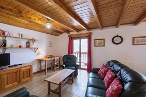 Un salon avec un plafond en bois, un canapé en cuir noir, une table en bois, un téléviseur posé sur un meuble et une fenêtre avec des rideaux rouges.
