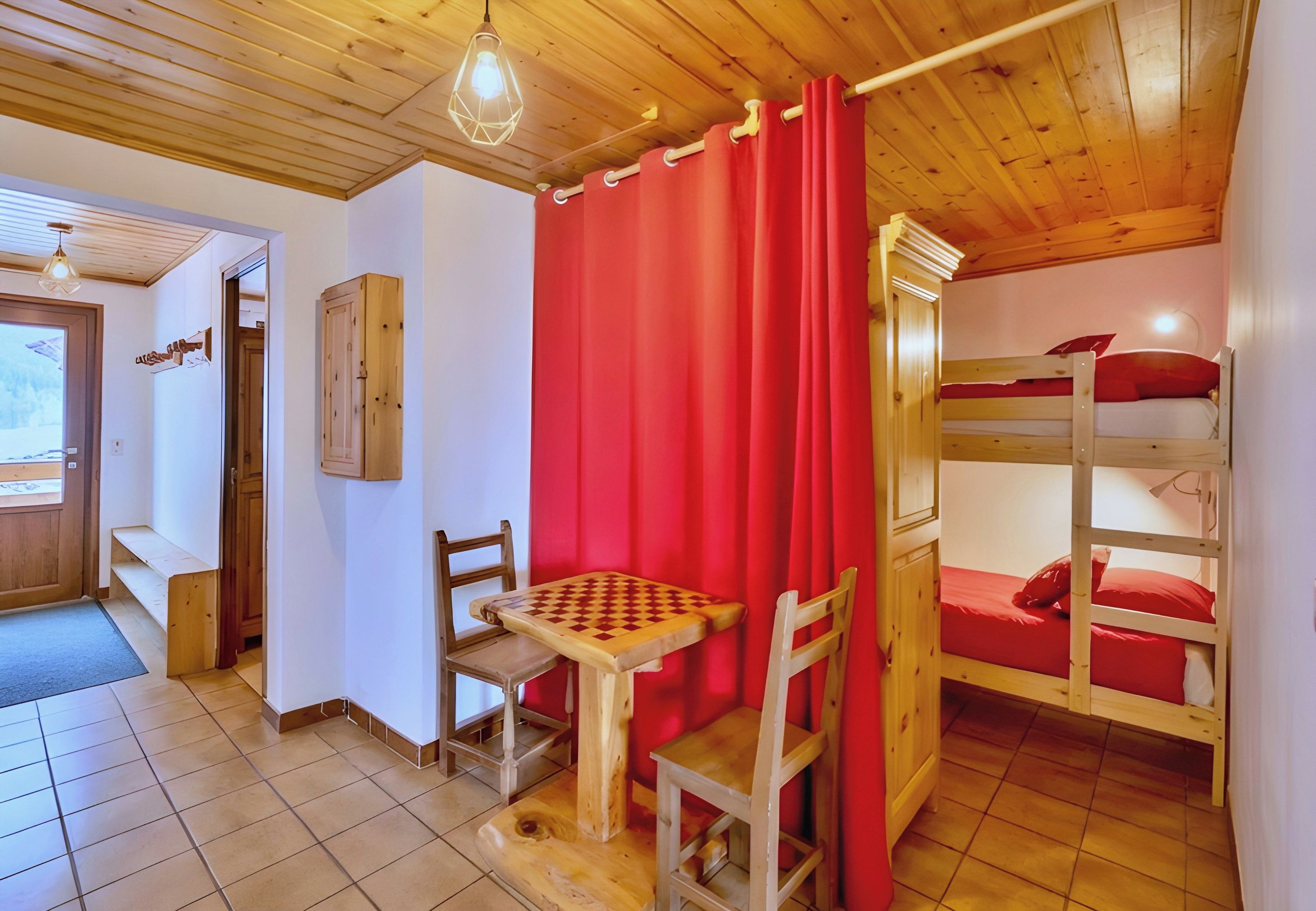 Une pièce avec un lit superposé en bois, une petite table avec des chaises, un rideau rouge et une armoire en bois.