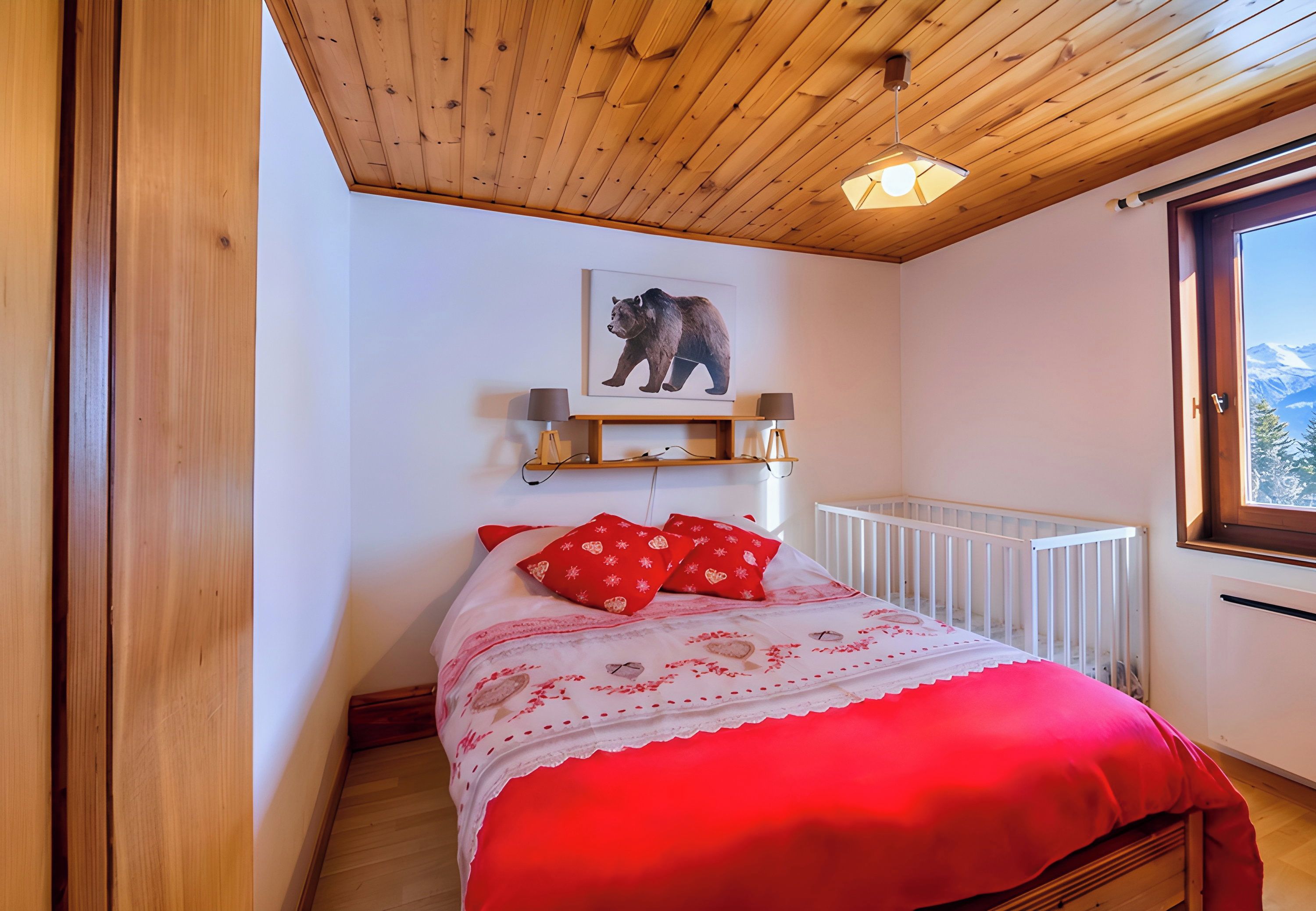 Une chambre avec un plafond en bois, un lit recouvert d’une couverture rouge, un tableau représentant un ours accroché au mur et une fenêtre donnant sur les montagnes.