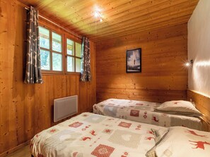 Quarto de cabana de madeira com duas camas, janela com cortinas, radiador e quadro na parede.