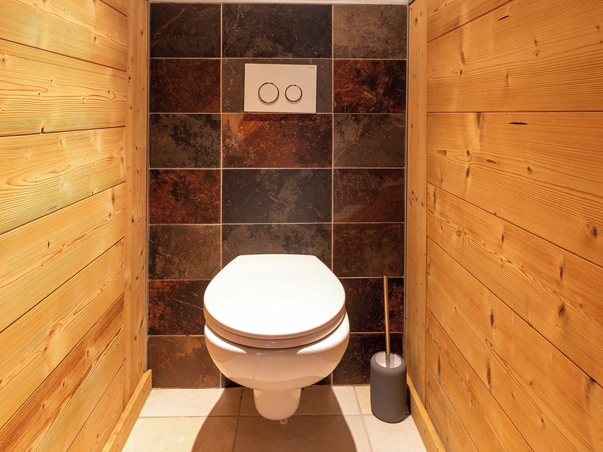 A small, wooden-paneled bathroom with a white toilet, a wall-mounted flush handle, and a toilet brush.