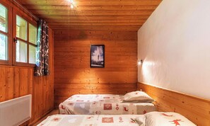 A wooden cabin room with two beds, a window with curtains, a framed picture on the wall, and a ceiling light.