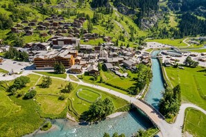 An aerial view of a mountain village with traditional wooden houses, a river, and a bridge.