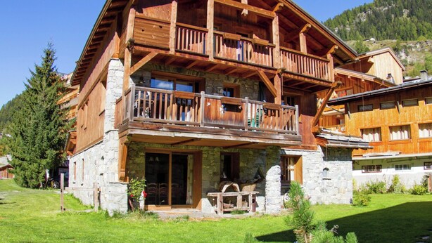 Un chalet de madera de dos pisos con balcones, cimientos de piedra y un paisaje montañoso de fondo.