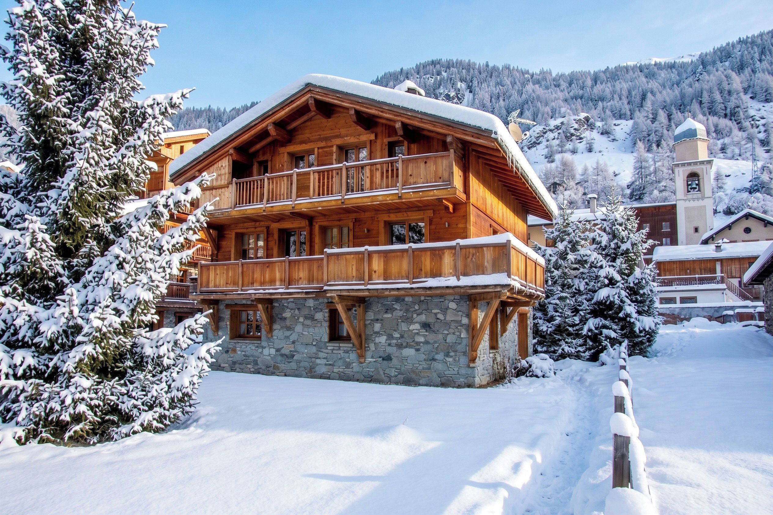 Ein mehrstöckiges Holzchalet mit Steinunterbau, umgeben von schneebedeckten Bäumen und einer Bergkulisse.