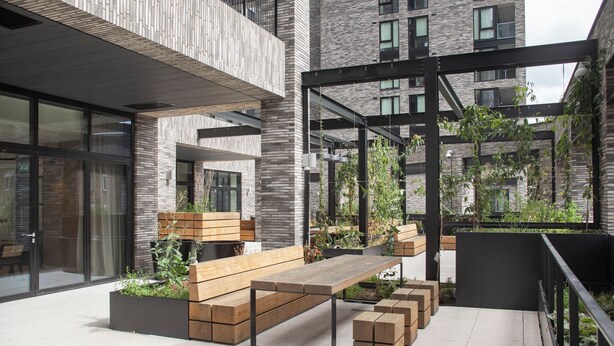 An outdoor seating area with wooden benches and planters, surrounded by modern buildings with large windows and a metal structure. - Enclave KX - Campus Accommodation (London)