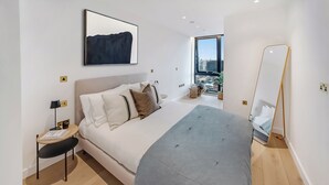 A modern bedroom with a large bed, a bedside table, a floor lamp, and a large window offering a city view. - Enclave KX - Campus Accommodation (London)