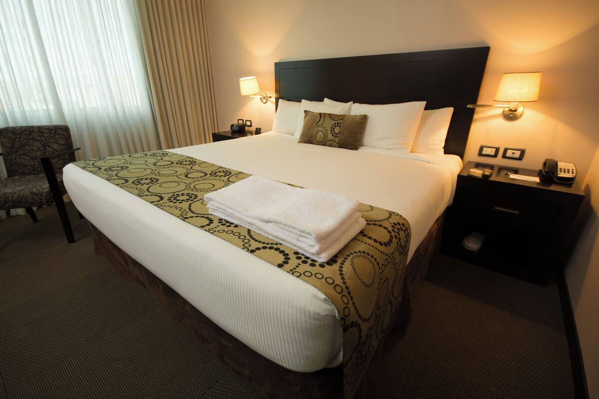 Una habitación de hotel con una cama grande, una silla, una mesita de noche con lámpara y una ventana con cortinas.