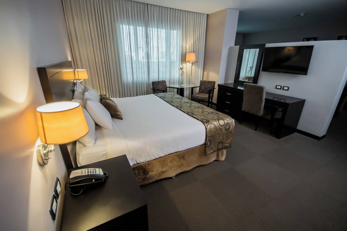 Una habitación de hotel con una cama grande, lámparas de noche, un escritorio con teléfono, televisor y una silla.