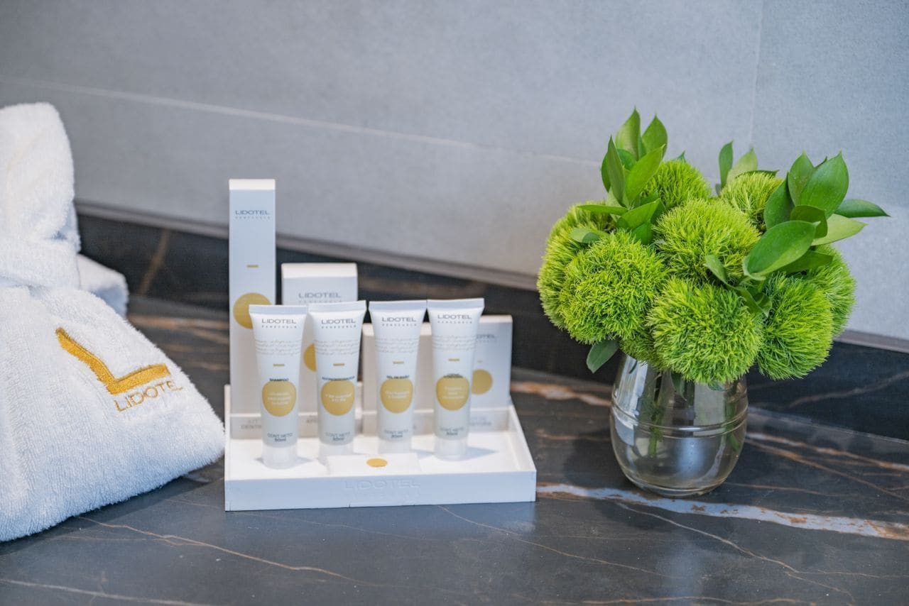 Une présentation de produits de soins de la peau LIDOEL sur un plateau blanc, une serviette blanche ornée du logo LIDOEL et un vase contenant des feuilles vertes.