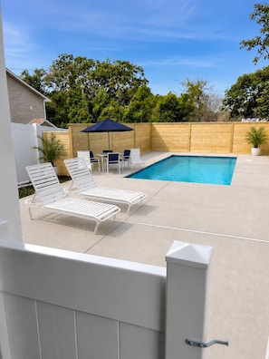 A backyard with a swimming pool, white lounge chairs, a table with chairs, and a wooden fence.