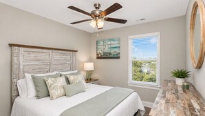 A bedroom with a bed, a ceiling fan, a nightstand with a lamp, a window with a view, and a wooden side table.
