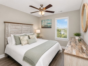 A bedroom with a bed, a ceiling fan, a nightstand with a lamp, a window with a view, and a wooden side table.