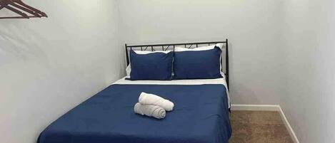 A small, neatly made bed with a blue bedspread and two pillows, a white towel folded on the bed, and a coat hanger on the wall.