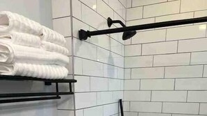 Un bagno con pareti piastrellate di bianco, una doccia con un tubo nero, un water e uno scaffale con asciugamani piegati.