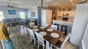 A spacious dining area with a wooden table and white chairs, a kitchen with white cabinets, and a gray sofa in the background. - Discover Your Dream Waterfront Getaway in Port Isabel, Texas! (Port Isabel)
