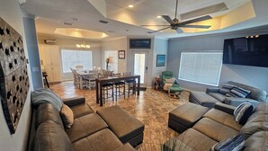 A spacious living room with a large sectional sofa, a dining area with a wooden table and chairs, a ceiling fan, and a view of the outdoors through the windows. - Discover Your Dream Waterfront Getaway in Port Isabel, Texas! (Port Isabel)