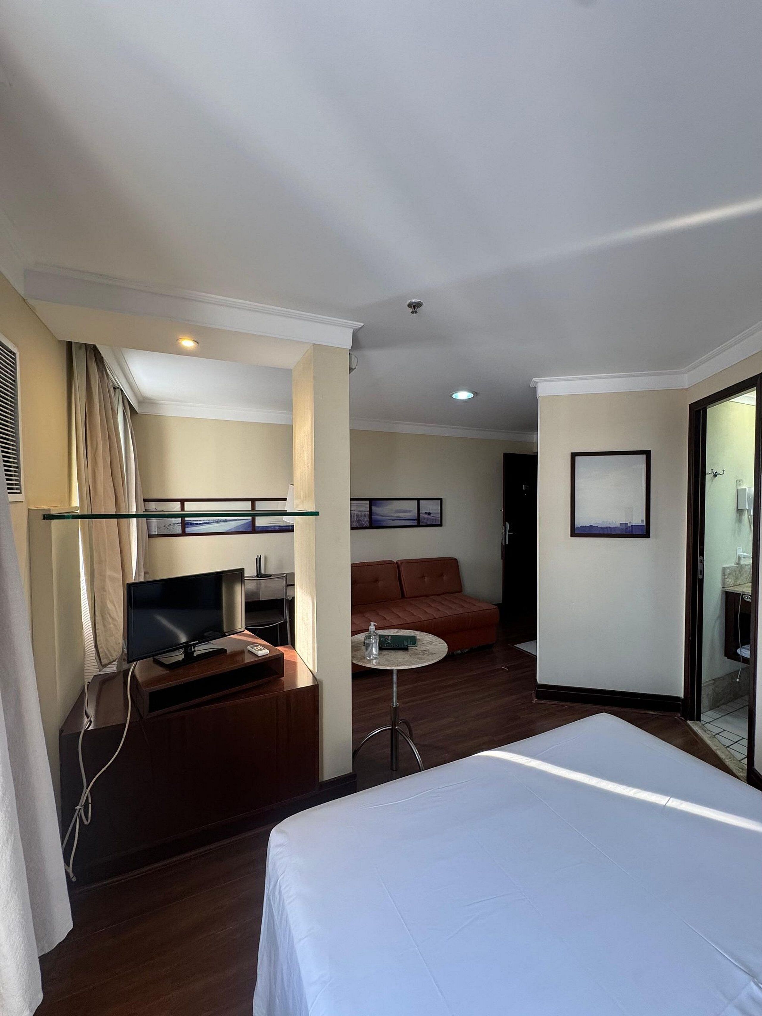 A hotel room with a bed, a TV, a small table, a sofa, and a glass-enclosed bathroom.