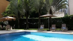 Ein Swimmingpool mit Liegestühlen und Sonnenschirmen, umgeben von Grünpflanzen und einem Gebäude im Hintergrund.