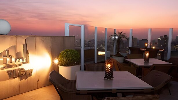 Eine Dachterrasse mit Sitzgelegenheiten und Tischen, einer Topfpflanze und Blick auf die Skyline der Stadt im Abenddämmerung.