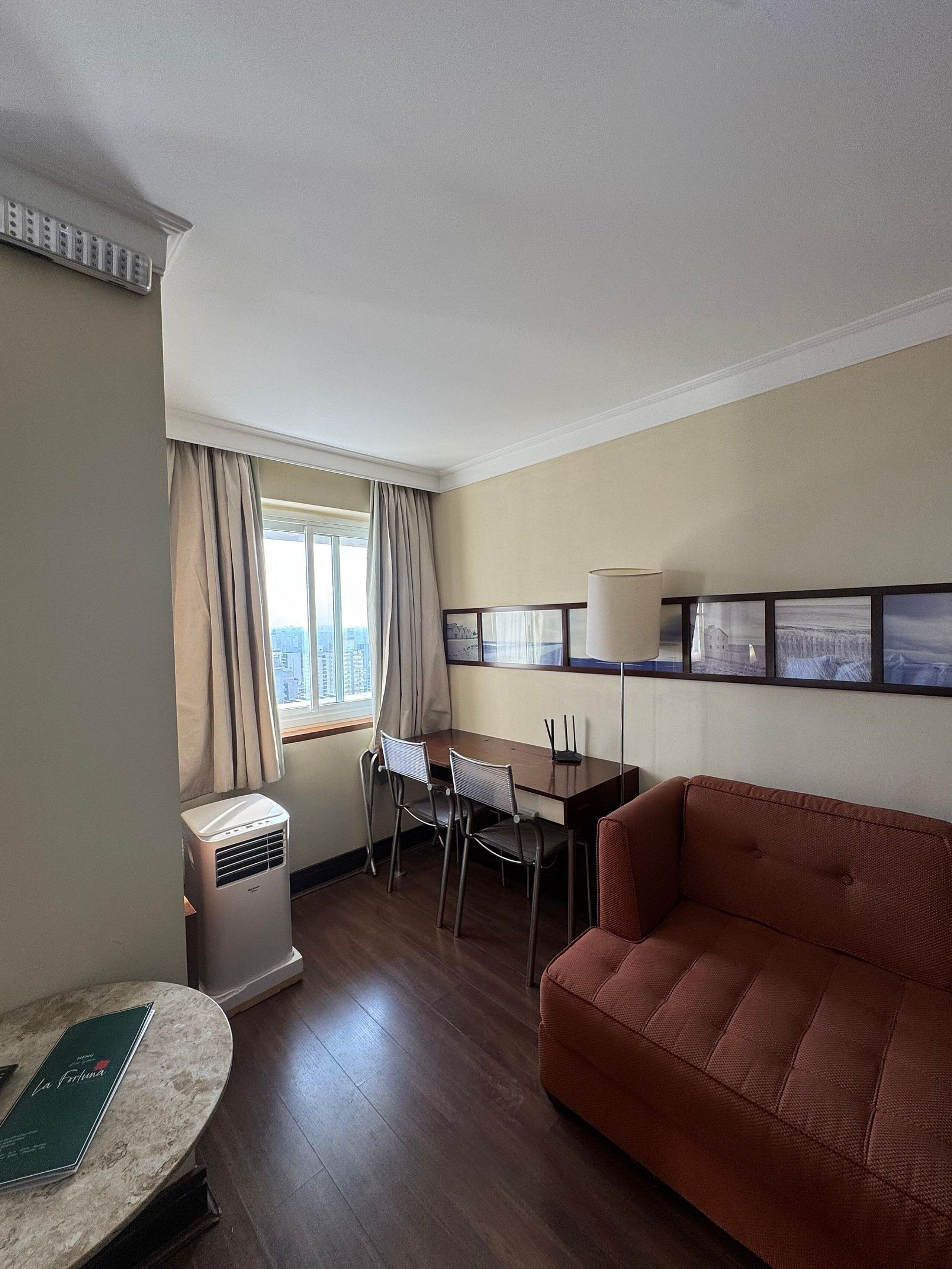 A hotel room with a wooden floor, a brown sofa, a dining table with chairs, a standing heater, and a window with curtains.