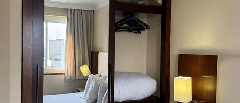 A hotel room with a wooden wardrobe, a bed, a bedside table with a lamp, and a telephone.