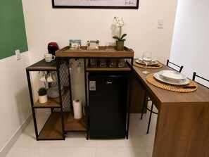 A compact kitchen with a refrigerator, a coffee machine, and a small shelf with kitchenware.