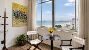 A room with a view of the beach, two chairs, a table with a vase of flowers, and a painting on the wall. - Amazing sea view in Copacabana (Rio de Janeiro)