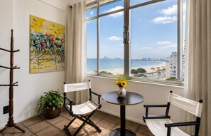 A room with a view of the beach, two chairs, a table with a vase of flowers, and a painting on the wall. - Amazing sea view in Copacabana (Rio de Janeiro)