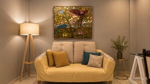 Een woonkamer met een beige bank, gekleurde kussens, een vloerlamp, een schilderij van vogels en een kleine tafel met een plant.