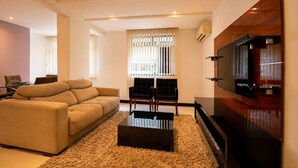 A modern living room with a sofa, a coffee table, a TV, and a window with blinds. - Confortável 3Qts: Vista Lateral Mar (Rio de Janeiro)