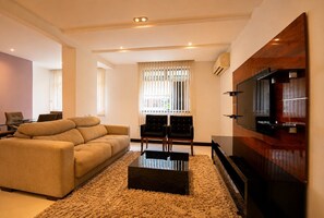 A modern living room with a sofa, a coffee table, a TV, and a window with blinds. - Confortável 3Qts: Vista Lateral Mar (Rio de Janeiro)
