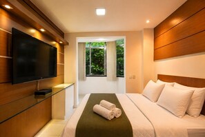 Habitación de hotel moderna con televisor de pantalla plana, un aparador de vidrio, cama con ropa blanca y vista a la vegetación por una ventana.