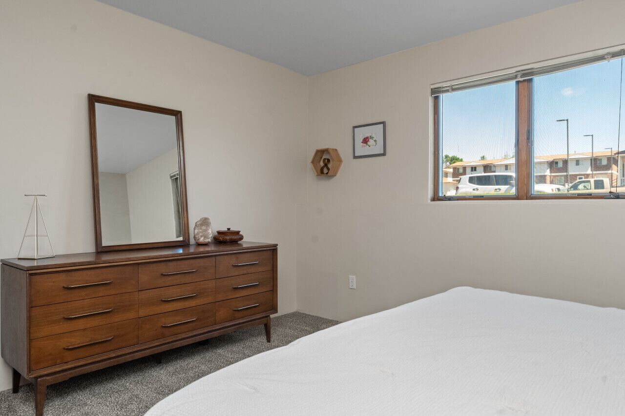 A bedroom with a wooden dresser, a large mirror, a framed picture, and a window with a view of parked cars.