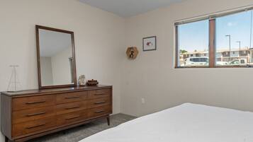 A bedroom with a wooden dresser, a large mirror, a framed picture, and a window with a view of parked cars.