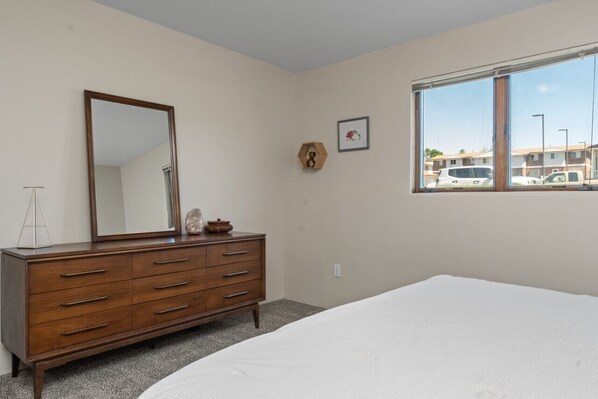 Une chambre à coucher avec une commode en bois, un grand miroir, un tableau encadré et une fenêtre donnant sur des voitures garées.