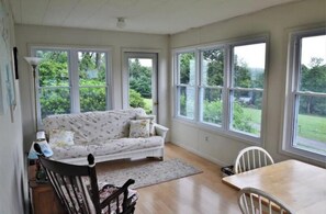 A cozy room with a sofa, a dining table, and chairs. Large windows offer a view of greenery outside.