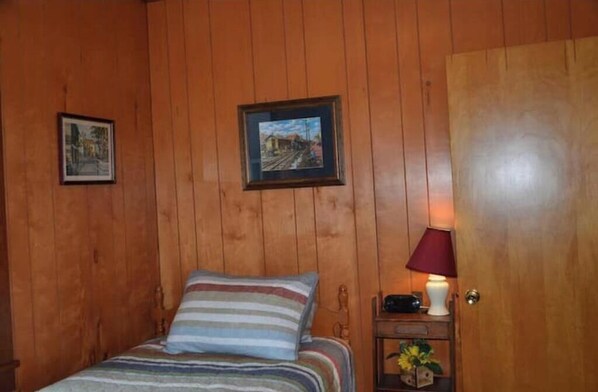 A wooden-paneled room with a bed, a nightstand with a lamp, and two framed pictures on the wall.
