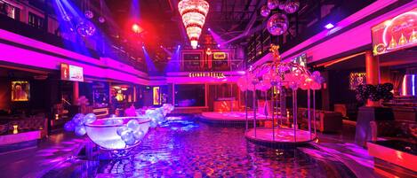 A luxurious indoor pool area with a central fountain, surrounded by seating and illuminated by vibrant lighting.