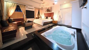 Une chambre d’hôtel moderne dotée d’un grand lit, d’un coin salon avec un canapé et des fauteuils, d’une baignoire et d’un téléviseur à écran plat fixé au mur.