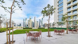 An outdoor seating area with tables and chairs, surrounded by trees and a view of a city skyline with modern skyscrapers.