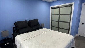 A bedroom with a bed, a nightstand, a dresser, a window with blinds, and a door.