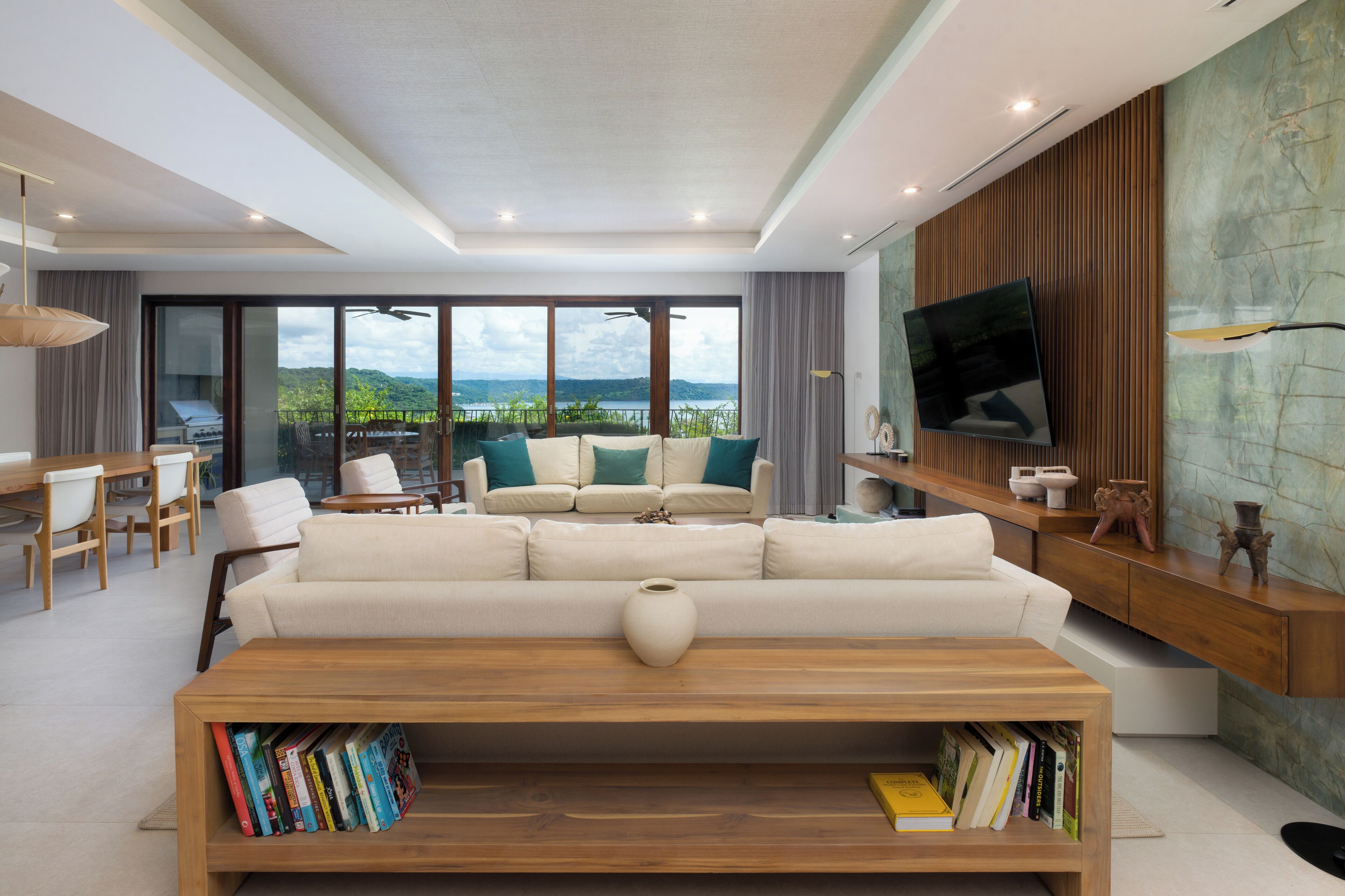 Un salon moderne avec un grand canapé, une table basse sur laquelle sont posés des livres, un téléviseur fixé au mur et une vue sur un lac et des montagnes, visible à travers de grandes fenêtres.