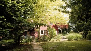 Et rødt hus med en stenkøbssti, der fører til hoveddøren, omgivet af frodig vegetation og træer.