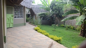 A stone pathway leading to a house with a thatched roof, surrounded by lush greenery and banana plants. - Hillstone Safari Lodge (Kimana)