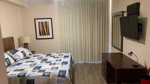 Ein Hotelzimmer mit Bett, Schreibtisch, Stuhl, Deckenventilator, Lampe, einem gerahmten Bild und einem Fenster mit Vorhängen.