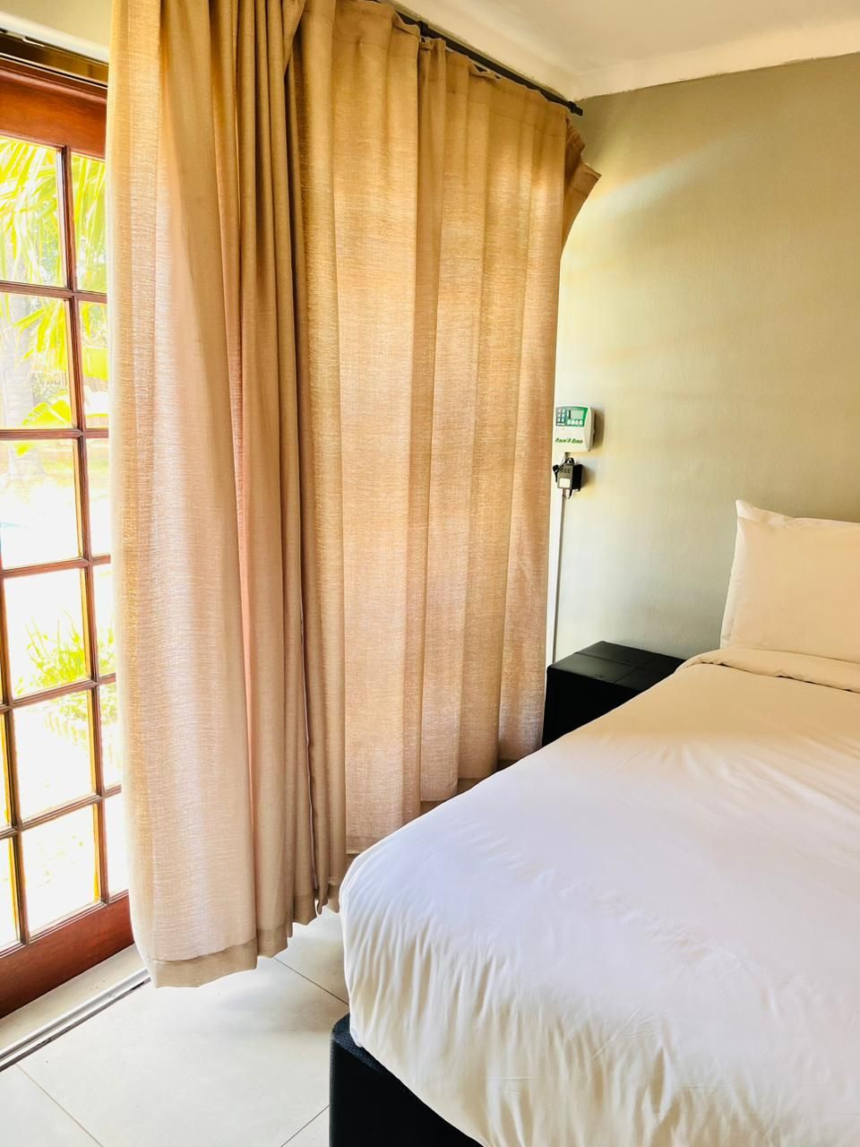 A hotel room with a bed, curtains, a nightstand, and a wall-mounted thermostat.