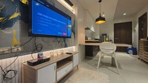 Una sala moderna con televisor de pantalla plana montado en una pared de mármol, mueble de madera para entretenimiento, una silla blanca y un área de cocina al fondo.